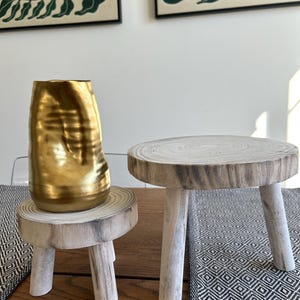 May include: A gold-colored, textured vase sits on a small, round wooden stool with three legs. Beside it is another similar stool. The stools are made of light-colored wood and have a natural, rustic appearance. The background includes framed botanical art.
