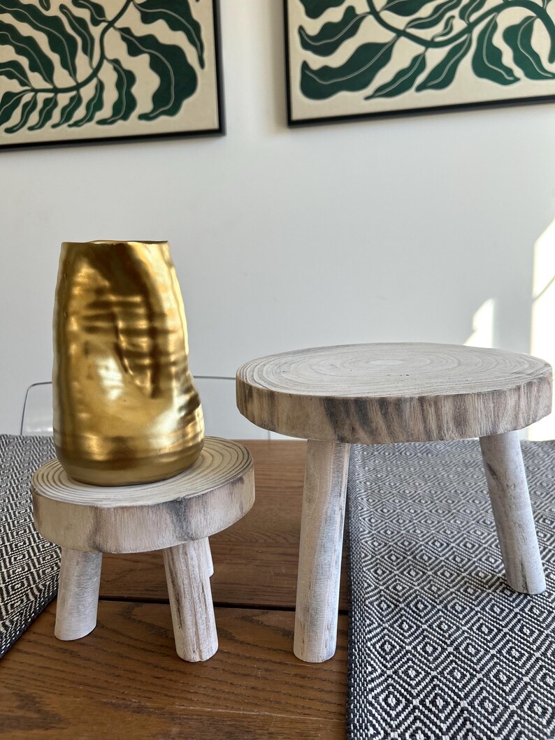 May include: A gold-colored, textured vase sits on a small, round wooden stool with three legs. Beside it is another similar stool. The stools are made of light-colored wood and have a natural, rustic appearance. The background includes framed botanical art.