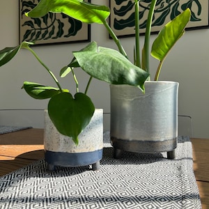 May include: Two cylindrical ceramic planters with three small feet each. The smaller planter has a speckled light grey top and a solid dark blue base. The larger planter features a smooth light grey finish with a dark blue bottom band.