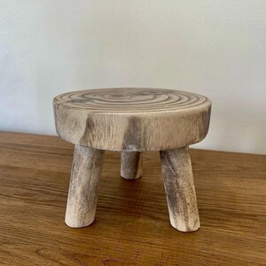 May include: A small, rustic wooden plant stand with a round top and three sturdy legs. The stand is made from a light-colored wood, showcasing the natural grain and texture. Perfect for displaying small plants or decorative items.