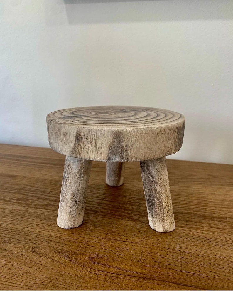 May include: A small, rustic wooden plant stand with a round top and three sturdy legs. The stand is made from a light-colored wood, showcasing the natural grain and texture. Perfect for displaying small plants or decorative items.