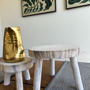 May include: Two small, rustic wooden stools with a natural wood grain finish. One stool is supporting a gold-colored vase. The stools have three legs each and are placed on a wooden table with a patterned runner.