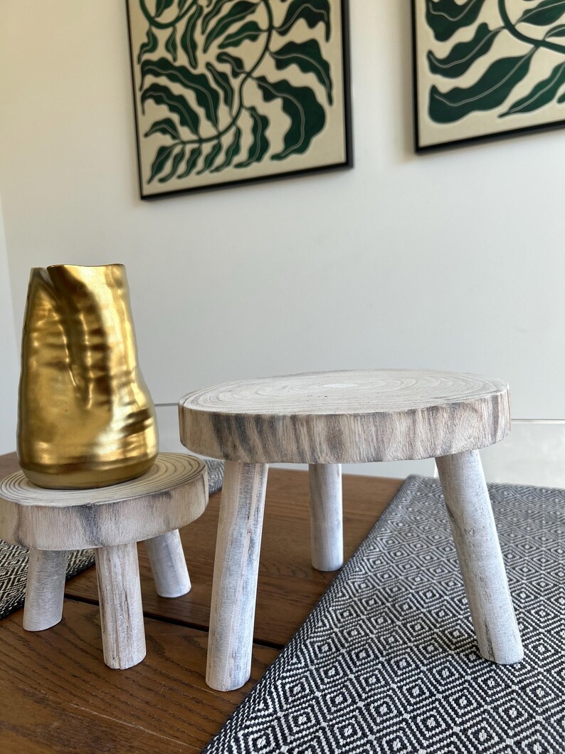May include: Two small, rustic wooden stools with a natural wood grain finish. One stool is supporting a gold-colored vase. The stools have three legs each and are placed on a wooden table with a patterned runner.