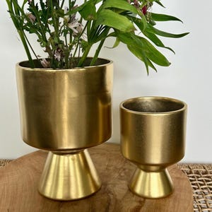 Puede incluir: Dos macetas de cerámica doradas con base acampanada. La maceta más grande es más alta y tiene una base más ancha. La maceta más pequeña es más baja y tiene una base más estrecha.