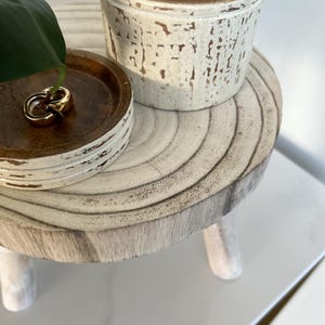 May include: A small, round, wooden side table with three legs. A small wooden tray with gold rings sits on the table. A white candle in a wooden container is also on the table. The table has a distressed white finish.