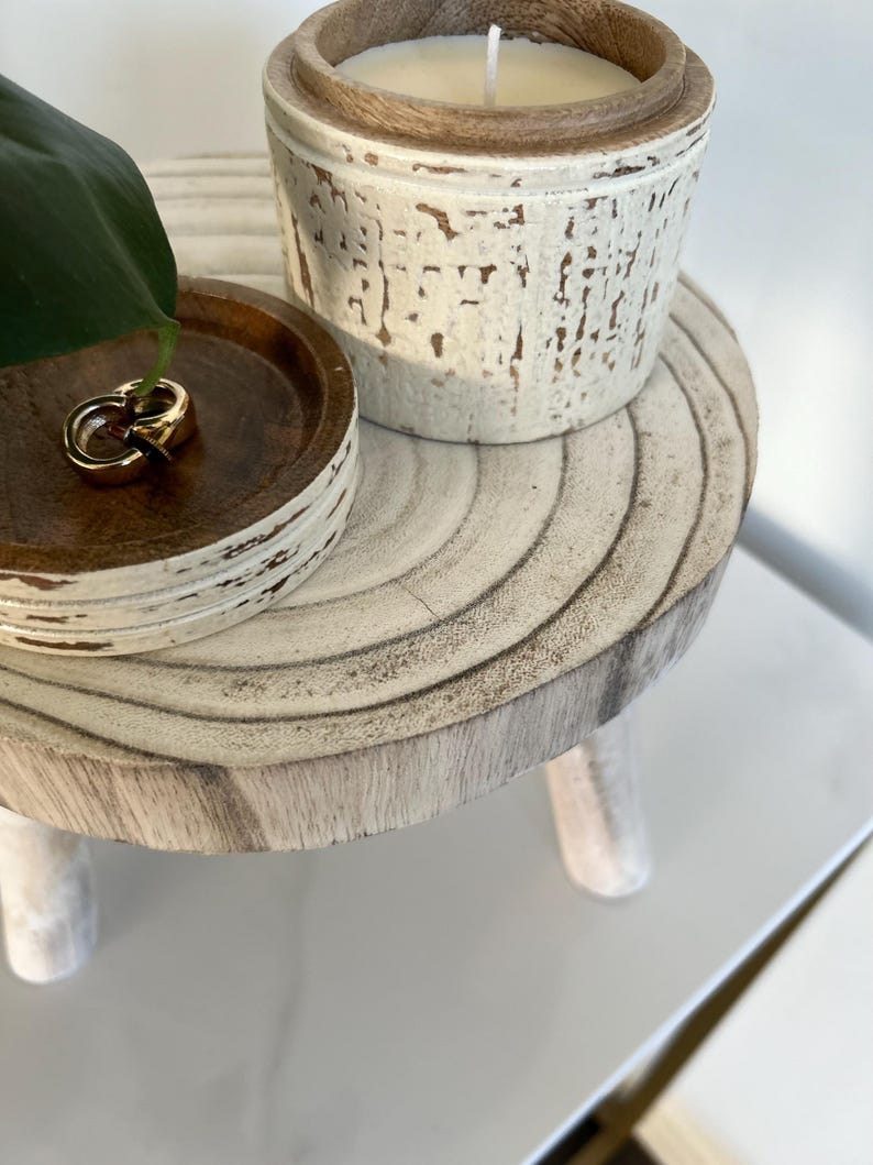 May include: A small, round, wooden side table with three legs. A small wooden tray with gold rings sits on the table. A white candle in a wooden container is also on the table. The table has a distressed white finish.