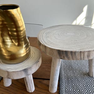 May include: Two small, rustic wooden plant stands with three legs each. One stand holds a gold-colored vase. The stands are made from a light-colored wood with visible grain. The stands are on a wooden surface, with a black and white patterned rug in the background.