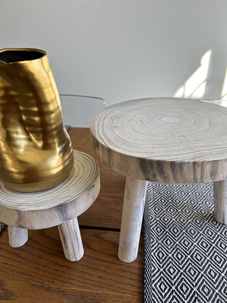 May include: Two small, rustic wooden plant stands with three legs each. One stand holds a gold-colored vase. The stands are made from a light-colored wood with visible grain. The stands are on a wooden surface, with a black and white patterned rug in the background.