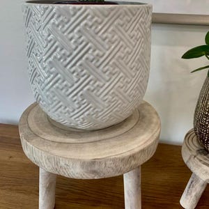 May include: A white ceramic planter with a geometric pattern sits atop a small, round, whitewashed wooden plant stand. The stand has three short legs. The planter contains a green plant.