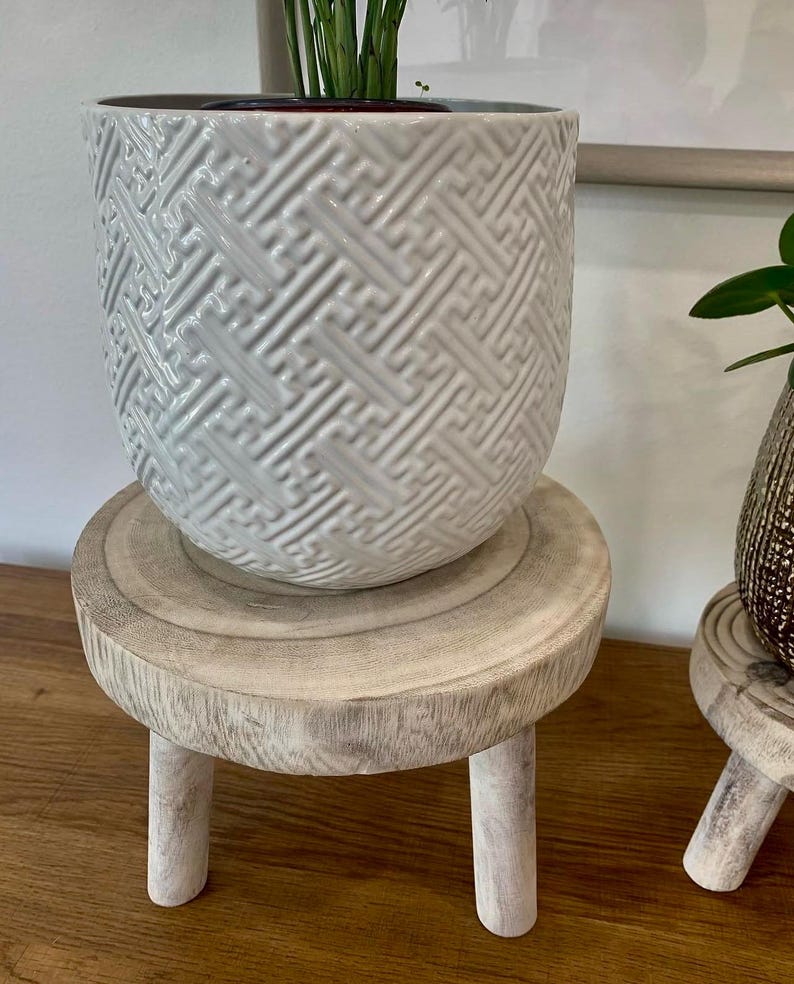 May include: A white ceramic planter with a geometric pattern sits atop a small, round, whitewashed wooden plant stand. The stand has three short legs. The planter contains a green plant.