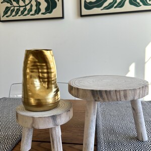 May include: A gold-colored, textured vase sits on a small, round wooden pedestal with three legs. Beside it is a similar pedestal. The pedestals are made of light-colored wood. Two framed botanical prints hang in the background.
