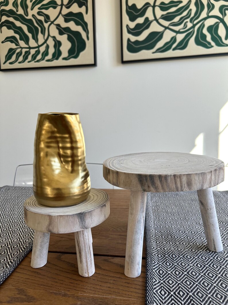 May include: A gold-colored, textured vase sits on a small, round wooden pedestal with three legs. Beside it is a similar pedestal. The pedestals are made of light-colored wood. Two framed botanical prints hang in the background.