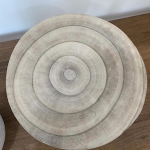 May include: A round, light-colored wooden table top with concentric rings, resembling tree rings. The table top is made of natural wood and has a rustic, organic aesthetic. The diameter is approximately 24 inches.