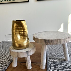 May include: A gold-colored, textured vase sits on a small, round wooden pedestal with three white-painted legs. Next to it is a similar pedestal. The pedestals are on a wooden table with a black and white patterned runner.