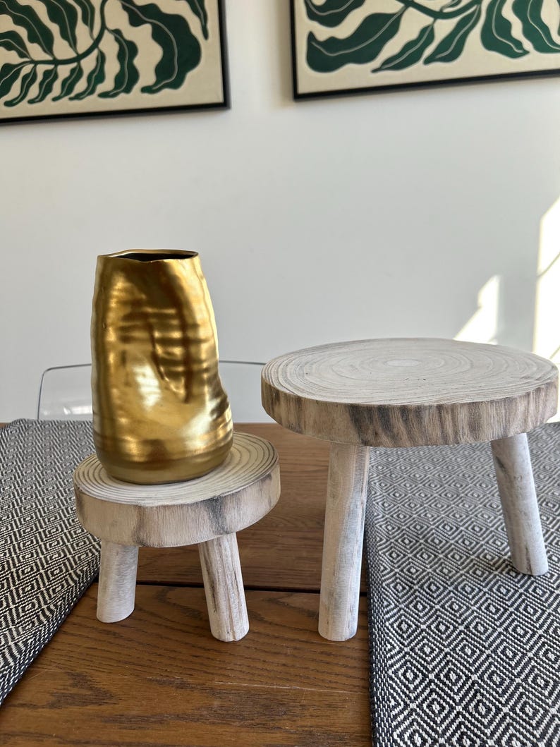May include: A gold-colored, textured vase sits on a small, round wooden pedestal with three white-painted legs. Next to it is a similar pedestal. The pedestals are on a wooden table with a black and white patterned runner.