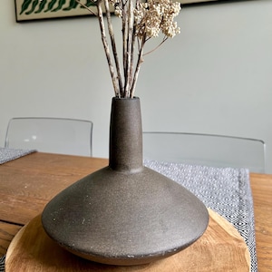 May include: A dark brown ceramic vase with a wide, shallow base and a narrow neck, holding dried white flowers. The vase sits on a wooden tray, with a patterned runner and a wooden table in the background. Decorative home decor.