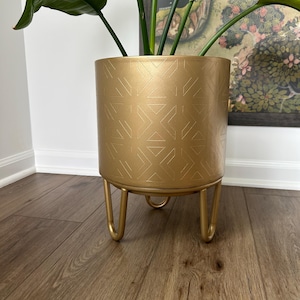 May include: A cylindrical gold metal planter featuring a repeating geometric pattern of etched lines. The planter is supported by three matching gold metal hairpin-style legs and contains a large green plant with broad, dark leaves.