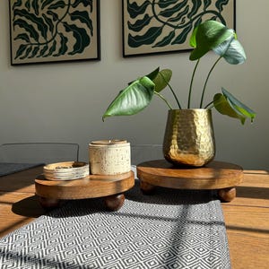 May include: A wooden table setting featuring two small, round wooden stands with decorative items. One stand holds a gold-colored hammered metal vase with a plant, and the other holds a small ceramic container and a stack of plates.