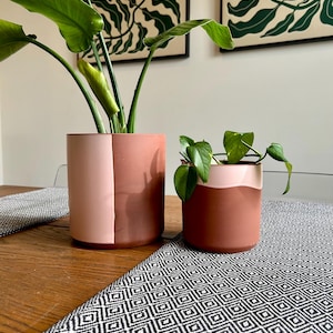 Può includere: Due vasi per piante in ceramica color terracotta, uno grande e uno piccolo, ciascuno con piante verdi. Il vaso più grande ha un design bicolore con una sezione rosa. I vasi sono su un tavolo di legno con un runner a fantasia bianco e nero.
