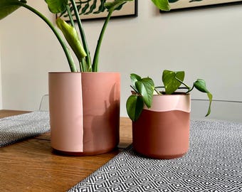 May include: Two terracotta-colored ceramic plant pots, one large and one small, each holding green plants. The larger pot has a two-tone design with a pink section. The pots sit on a wooden table with a black and white patterned runner.
