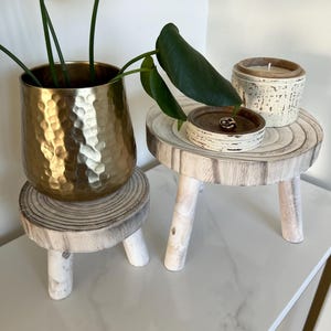 May include: A gold hammered metal planter with a green plant sits on a small, white-washed wooden stool. A small wooden tray and a candle in a textured ceramic container are on a similar stool. The items are arranged on a white surface.