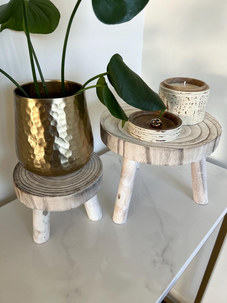 May include: A gold hammered metal planter with a green plant sits on a small, white-washed wooden stool. A small wooden tray and a candle in a textured ceramic container are on a similar stool. The items are arranged on a white surface.