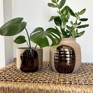 May include: Two ceramic planters with a brown glaze and a textured design. The larger planter is holding a green plant with small leaves. The smaller planter is holding a green plant with large leaves. Both planters are sitting on a woven wicker surface.