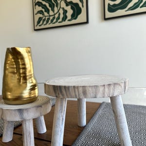 May include: Two small, rustic wooden stools with three legs each. One stool is topped with a gold-colored vase. The stools are on a wooden surface with a black and white patterned runner.