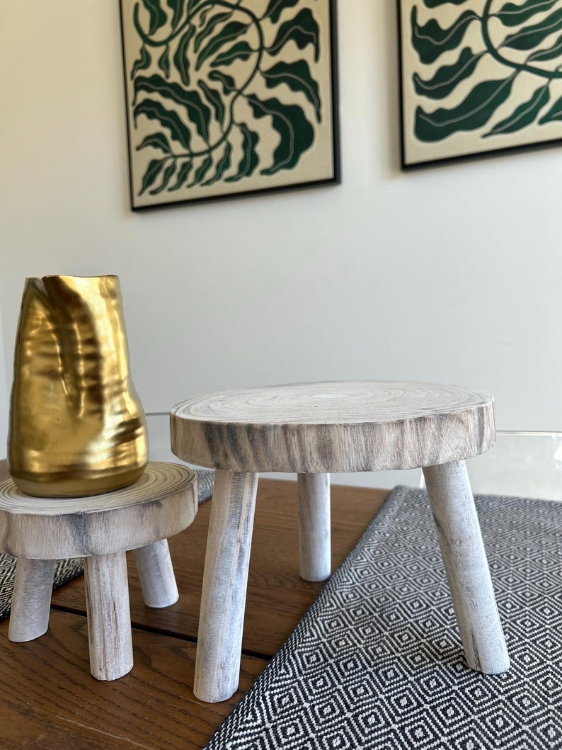 May include: Two small, rustic wooden stools with three legs each. One stool is topped with a gold-colored vase. The stools are on a wooden surface with a black and white patterned runner.