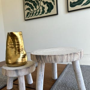 May include: Two small, rustic wooden stools with three legs each. One stool has a gold-colored vase on top. The stools are made of light-colored wood with a natural grain pattern. The legs are painted white.