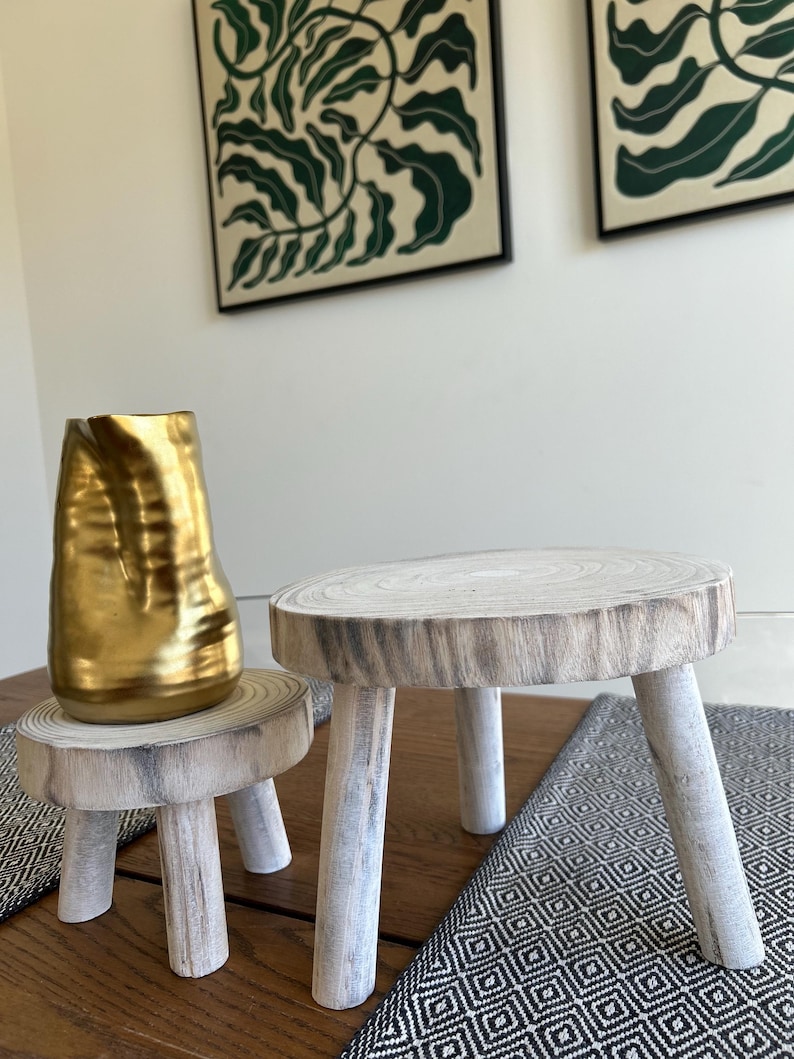May include: Two small, rustic wooden stools with three legs each. One stool has a gold-colored vase on top. The stools are made of light-colored wood with a natural grain pattern. The legs are painted white.