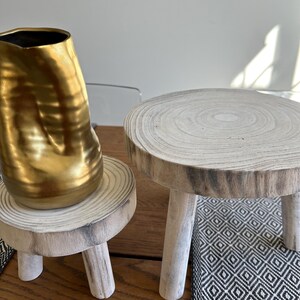 May include: A gold-colored, textured vase sits on a small, round wooden plant stand with three legs. Another similar stand is next to it. The stands are made of light-colored wood and are on a wooden surface.