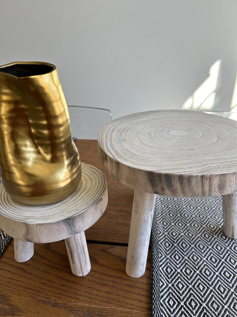 May include: A gold-colored, textured vase sits on a small, round wooden plant stand with three legs. Another similar stand is next to it. The stands are made of light-colored wood and are on a wooden surface.