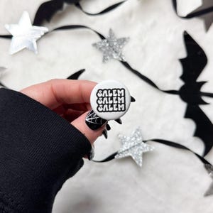 May include: A white button with the word "SALEM" stacked three times in a black, stylized font. The button is held against a backdrop of silver glitter stars and black bat and ribbon decorations, creating a Halloween theme.