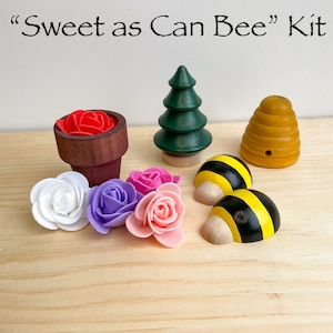 Puede incluir: Una colmena de madera, dos abejas negras y amarillas, un peque&ntilde;o &aacute;rbol verde, una maceta con una flor roja y cuatro flores artificiales de color rosa, blanco y morado. El texto "Sweet as Can Bee" Kit est&aacute; escrito encima de los art&iacute;culos.