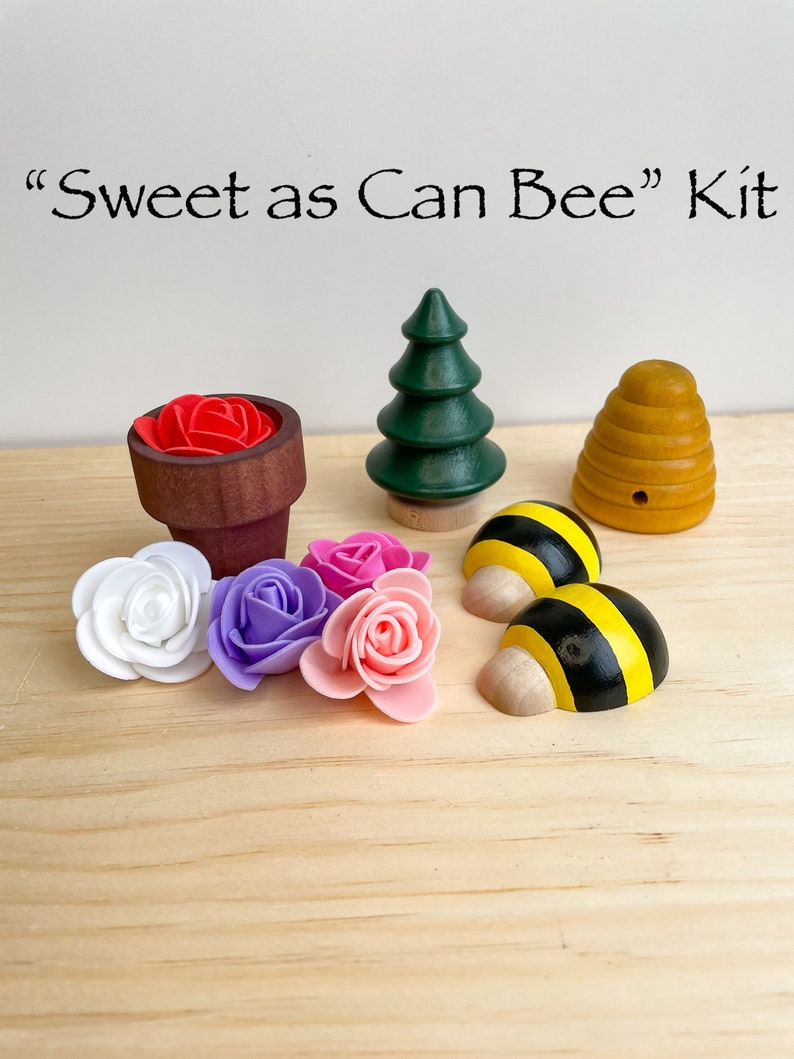 Puede incluir: Una colmena de madera, dos abejas negras y amarillas, un peque&ntilde;o &aacute;rbol verde, una maceta con una flor roja y cuatro flores artificiales de color rosa, blanco y morado. El texto "Sweet as Can Bee" Kit est&aacute; escrito encima de los art&iacute;culos.