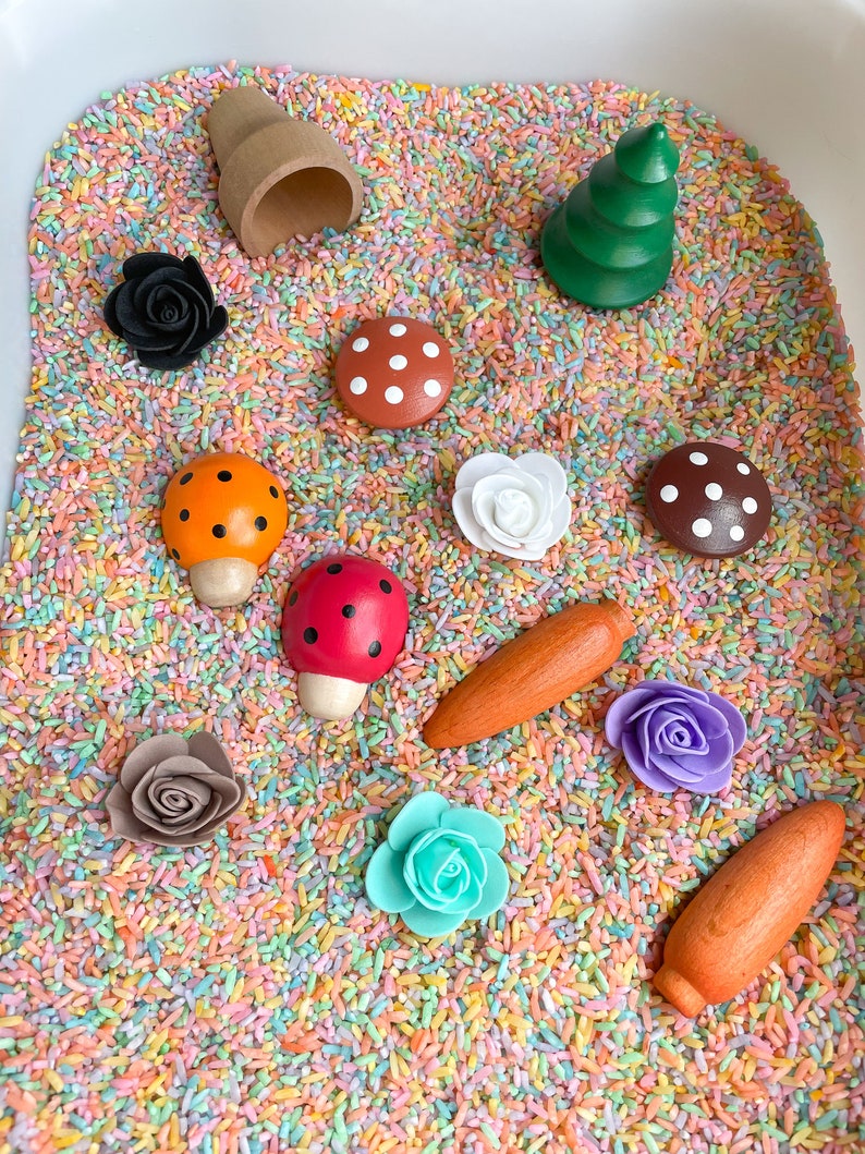 Puede incluir: Un contenedor sensorial lleno de arroz de colores y varios juguetes de madera, incluyendo un &aacute;rbol peque&ntilde;o, setas, mariquitas, flores y una maceta.