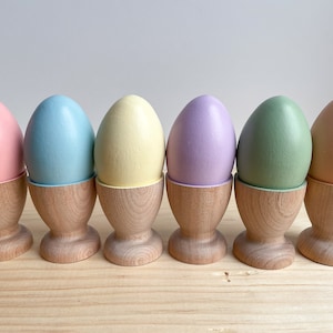 May include: Six pastel colored eggs in wooden egg cups. The eggs are pink, light blue, yellow, lavender, green, and peach.