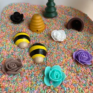 Puede incluir: Un contenedor sensorial lleno de arroz de colores y varios juguetes de madera y pl&aacute;stico, incluyendo una colmena, una abeja, un &aacute;rbol, una flor y un hongo marr&oacute;n.