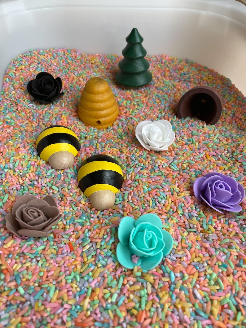 Puede incluir: Un contenedor sensorial lleno de arroz de colores y varios juguetes de madera y pl&aacute;stico, incluyendo una colmena, una abeja, un &aacute;rbol, una flor y un hongo marr&oacute;n.