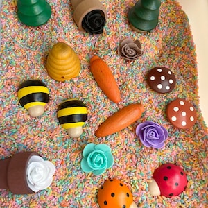 Puede incluir: Un contenedor sensorial lleno de arroz de colores y varios juguetes de madera, incluyendo &aacute;rboles, setas, flores, una abeja, una colmena y una mariquita.
