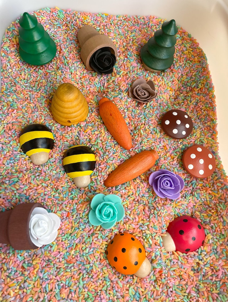 Puede incluir: Un contenedor sensorial lleno de arroz de colores y varios juguetes de madera, incluyendo &aacute;rboles, setas, flores, una abeja, una colmena y una mariquita.