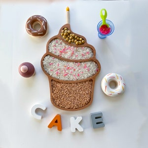 以下が含まれることがあります： ろうそくが置かれた木製カップケーキトレイ。白いご飯、ピンクのスプリンクル、茶色いご飯で満たされています。トレイは「CAKE」と綴られた木製文字と、茶色と白のドーナツ2つで囲まれています。
