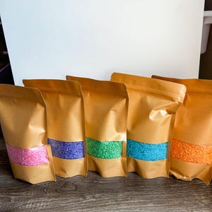 Peut inclure: Cinq sacs en papier brun avec des fermetures refermables, chacun rempli d'une couleur de riz différente. Les couleurs sont rose, violet, vert, bleu et orange.