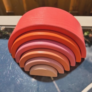 May include: A wooden rainbow stacking toy with six arches in shades of red, orange, pink, and beige. The arches are arranged in descending size, with the largest arch at the top and the smallest arch at the bottom.
