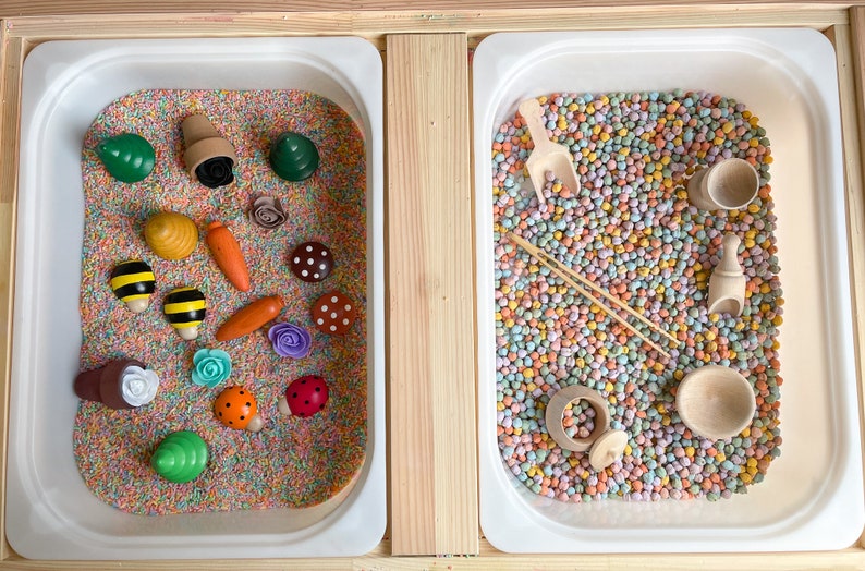 Puede incluir: Dos bandejas blancas llenas de materiales sensoriales coloridos. La bandeja de la izquierda tiene una variedad de peque&ntilde;os juguetes de madera, incluyendo flores, mariquitas, abejas y zanahorias. La bandeja de la derecha tiene una pala, un palo de madera y un peque&ntilde;o cuenco de madera.