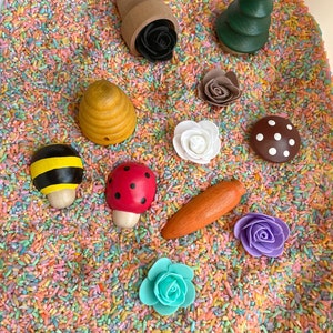 Puede incluir: Un contenedor sensorial lleno de arroz de colores y juguetes de madera, incluyendo una abeja, una mariquita, un hongo, una zanahoria, flores, un &aacute;rbol y una colmena.