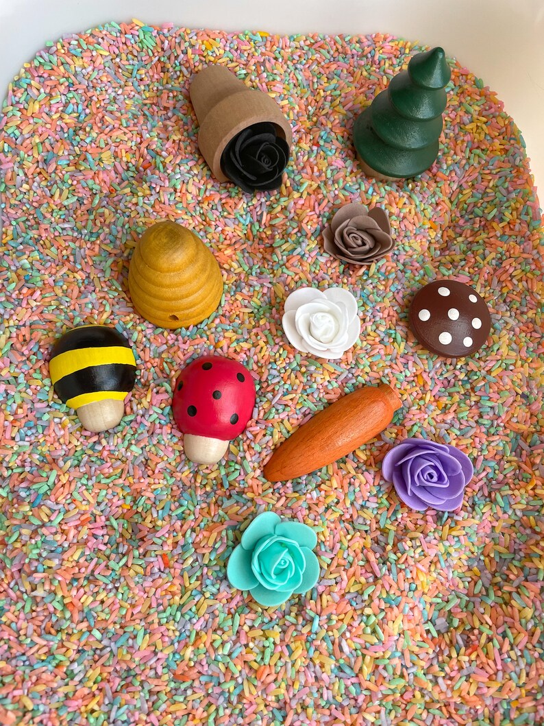 Puede incluir: Un contenedor sensorial lleno de arroz de colores y juguetes de madera, incluyendo una abeja, una mariquita, un hongo, una zanahoria, flores, un &aacute;rbol y una colmena.