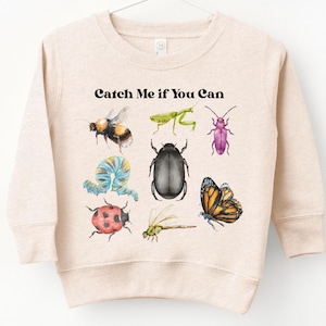 May include: Beige sweatshirt featuring colorful insect illustrations, including a bee, grasshopper, and butterfly. The text "Catch Me If You Can" is printed above the insects. The sweatshirt is hanging on a white hanger.
