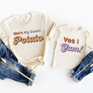 Pu&ograve; includere: Due magliette color crema con le frasi "She's My Sweet Potato" e "Yes I Yam!" in lettering in stile retr&ograve;. Le magliette sono abbinate a jeans blu ed erba secca decorativa, suggerendo un outfit coordinato per una famiglia o una coppia.
